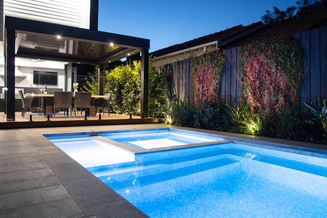 Evening view of illuminated pool and spa beside outdoor dining and greenery