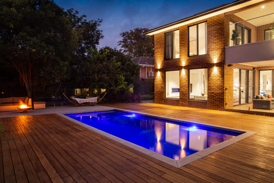 Backyard pool lit up at night with deck, fire pit and hammock