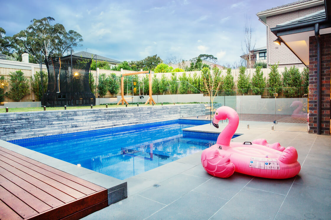 Blue tiled pool with pink flamingo float, play area and trampoline