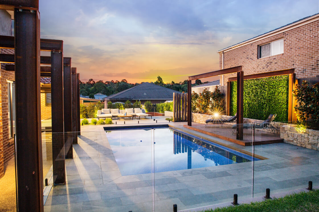 Pool at sunset with glass fence, stone paving and covered entertaining areas